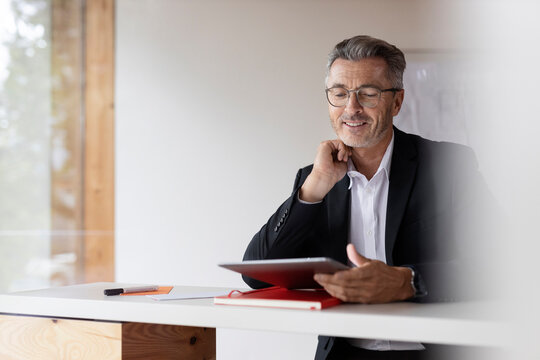 Smiling businessman with hand on chin using digital tablet at home