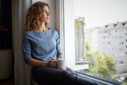 Woman with coffee cup looking through window while sitting on window sill at home