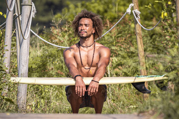 Muscular man with surfboard crouching against plants