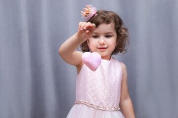 A pink soft toy in the shape of a heart in the hands of a 4-5 year old girl. Valentine's Day for Valentine's Day. Close-up portrait on gray background