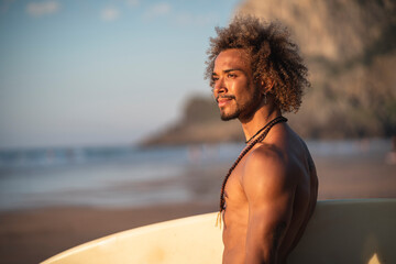 Smiling shirtless young man looking away while standing with surfboard at beach during sunset