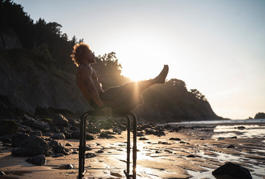 Young man exercising on parallel bars at beach against sky
