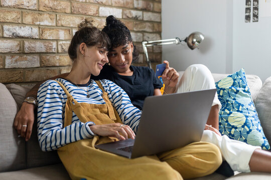 Woman with laptop holding credit card while sitting by girlfriend at home