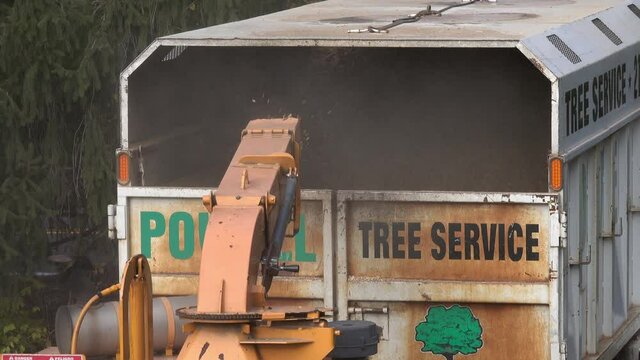 Wood chips are expelled from the chute of a wood chipper into a truck bed.  Dust rises from back of truck.