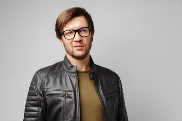 Portrait of a young man in glasses wearing black leather jacket on grey background