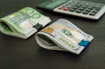 calculator, 100 euros and 100 dollars squeezed by a metal clip on a black wooden background.