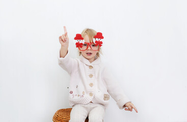 a little girl in funny Christmas glasses is sitting against a white wall.