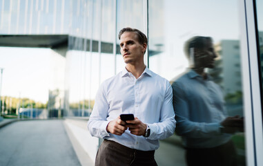 Contemplative male in white shirt standing near urban building thinking about financial news received on smartphone device, millennial businessman using cellular technology in metropolitan downtown