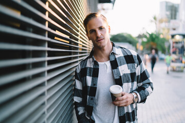 Half length portrait of pensive caucasian man 20s in trendy casual wear holding coffee cup standing on urban setting background, serious male hipster resting during weekend free time on street