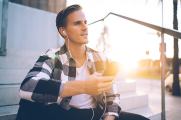 Handsome caucasian man in casual wear enjoying free time on sunny day listening audiobook via smartphone for education, young 20s male millennial in earphones sitting in town using technology