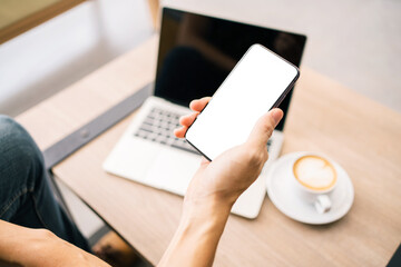 Cell phone mockup image with empty white screen. Male hand holding texting using mobile phone over the desk with laptop and coffee cup on the table in background. empty space for advertising text.