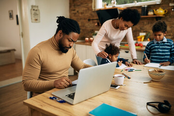 African American father examining reports while working on laptop at home.