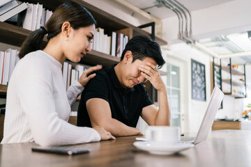 The sad unhappy handsome man sitting in front of a laptop, holding his forehead while having a headache with a woman cheering up by touching his shoulder.