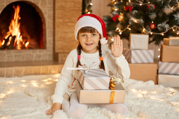 Fototapeta na wymiar happy child girl sitting near Christmas tree on Christmas Eve on floor and waving hand to camera, posing with stack of gift boxes, kid wearing casual clothing and red santa hat.