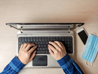 Fototapeta premium A man is typing on a laptop while sitting on the couch. The medical mask is nearby.