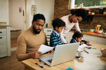 Black working father analyzing paperwork while mother is assisting their kids with homework at home.
