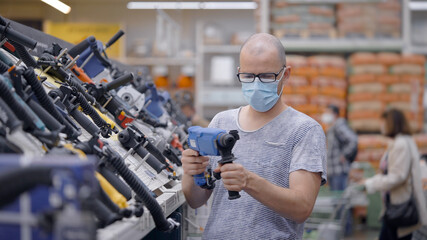 A man woker in a protective mask and wearing glasses chooses a electric drill at an tools store