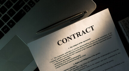 Top view of laptop computer, contract, planner and silver pen on wooden table in office. Warm sunlight.