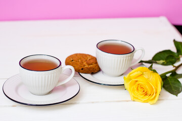 Two cups of tea and oatmeal cookies on a white wooden table and pink background. Breakfast. Yellow rose.