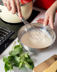 Close up of a woman is hand using a whisk to stir cheese sauce in a black saucepan, blurred background