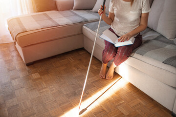 Young blind woman reading book written in Braille at home with white cane . Blind person reading book written in Braille. Young blind woman reading a braille book while studying and sitting at home