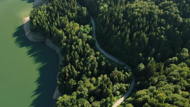 Aerial view of car driving through the fores and the lake on the side. Beautiful mountain road. Driving on the mountain road. Road trip. Mountain forest lake landscape. Car driving through pine forest