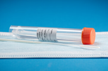 Swab (sample collection) with transport vial with orange cap, transparent with adhesive for data, on face mask with blue background.