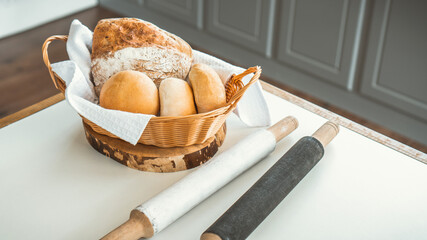 A basket containing fresh crisp bread. Nearby is a rolling pin for dough on a white table.