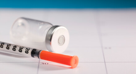 syringe with orange cannula and transparent vial with aluminum closure, on calendar, blue background