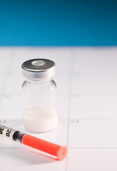 transparent vaccine vial with aluminum closure and syringe with orange cannula, on calendar, blue...