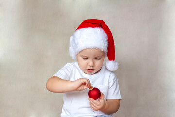 Portrait of a cute little boy in a white t-shirt and Santa Claus hat. A boy admires a red Christmas ball. Children's emotions. Christmas and new year.