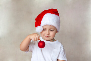 Portrait of a cute little boy in a white t-shirt and Santa Claus hat. A boy admires a red Christmas ball. Children's emotions. Christmas and new year.