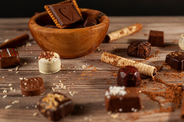 A bowl of assorted chocolates and chocolate biscuits on wooden background with cacao powder cinnamon sticks, pirouettes and coconut sprinkles creating an organic look. For Valentines day and christmas