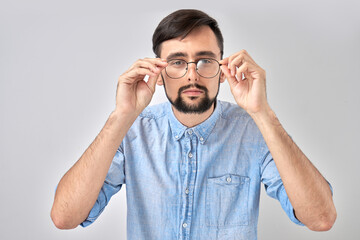 Studio shot caucasian bearded man with poor eyesight holds hands on the rim of glasses and squints, trying to see something, looking at the camera isolated on blue background