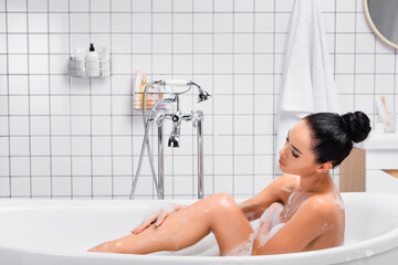 Obraz premium Young woman touching leg while bathing with foam in modern bathroom