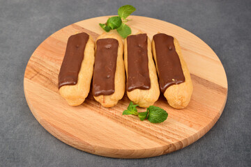 Four eclairs lie on a wooden cutting board. Several mint leaves are placed nearby