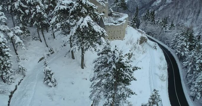 Aerial drone overhead view of medieval Predil fortress in Slovenia. Paved countryside road between forest. Cold winter in Alpine valley. Beautiful landscape covered with snow. Ascending