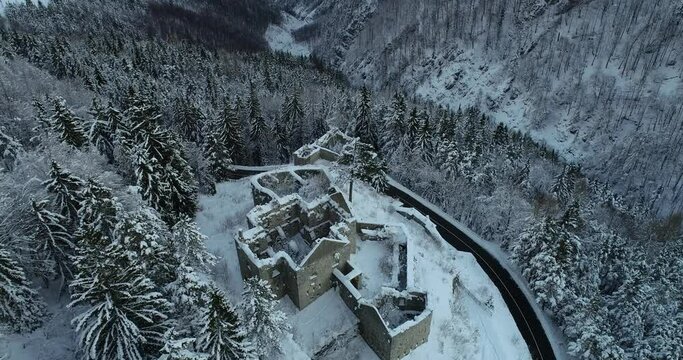 Aerial drone overhead view of medieval Predil fortress in Slovenia. Paved countryside road between forest. Cold winter in Alpine valley. Beautiful landscape covered with snow. Forward moving, tilt