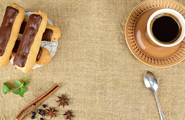 Top view of chocolate eclairs, hot coffee mug and teaspoon. Nearby are mint leaves, a cinnamon stick and star anise