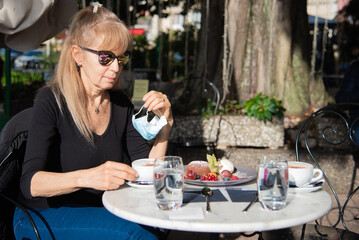 Blonde woman taking off her surgical mask to have a coffee and cake in an open-air cafe shop. Concept of new normality