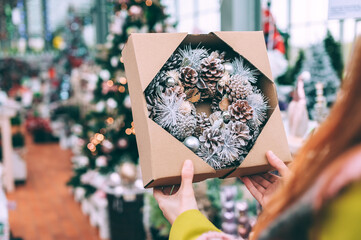 Naklejka na ściany i meble New Year's wreath in a packing box in the hands of a girl. Against the background of a Christmas tree in a supermarket.