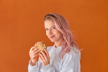 A young woman with pink hair looks at the camera and holds a Burger in her hand. Orange background. The concept of junk food.