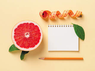 Half of ripe juicy grapefruit, roll of measuring tape, spring notepad and pensil on a pastel yellow background. Slimming diet, body weight control and healthy eating. Copy space.