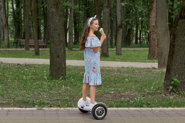 A girl rides a gyro scooter and eats ice cream in the Park. Beautiful blue dress. Summer green Park.