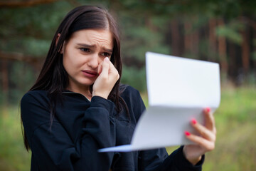 young woman is shocked and crying to see message in a letter on paper. outdoors