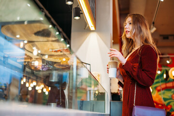 young beautiful woman with cup of coffee in her hands is standing at window, choosing a cake