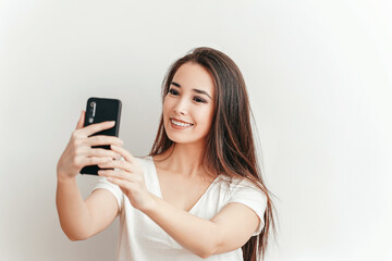 Beautiful Asian smiling girl takes selfie on mobile phone on white background in white T-shirt with beautiful smile