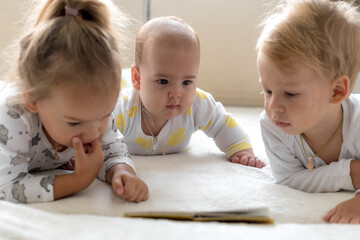 Three Little smiling kids play together on soft white bed at home. Brother and siste read book fairy tales with baby. Children meeting new born sibling.Toddlers laugh and bond. funny.Copy space