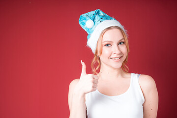 Cute blond woman smiling and showing thumb up in white tank top and blue christmas hat isolated on red background.