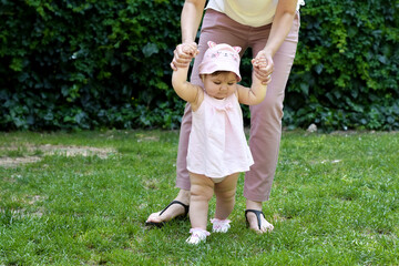 Cute  adorable baby girl taking first steps with mother's help (holding mums hands) in park (garden). Happy family moments. Family time. Happy childhood. Mother love, mother care and happy motherhood 